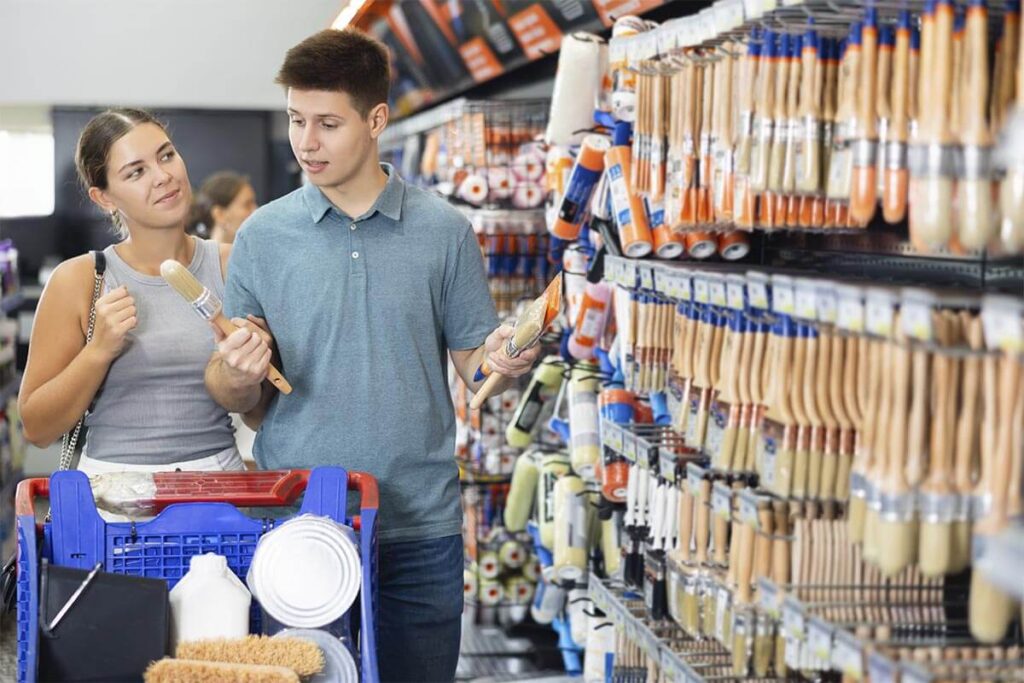 Casal fazendo compras em uma loja de materiais de construção, escolhendo pincéis na seção de ferramentas de pintura.