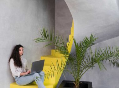 Mulher no laptop em ambiente com escada amarela e planta, com cores na decoração que inspiram home office