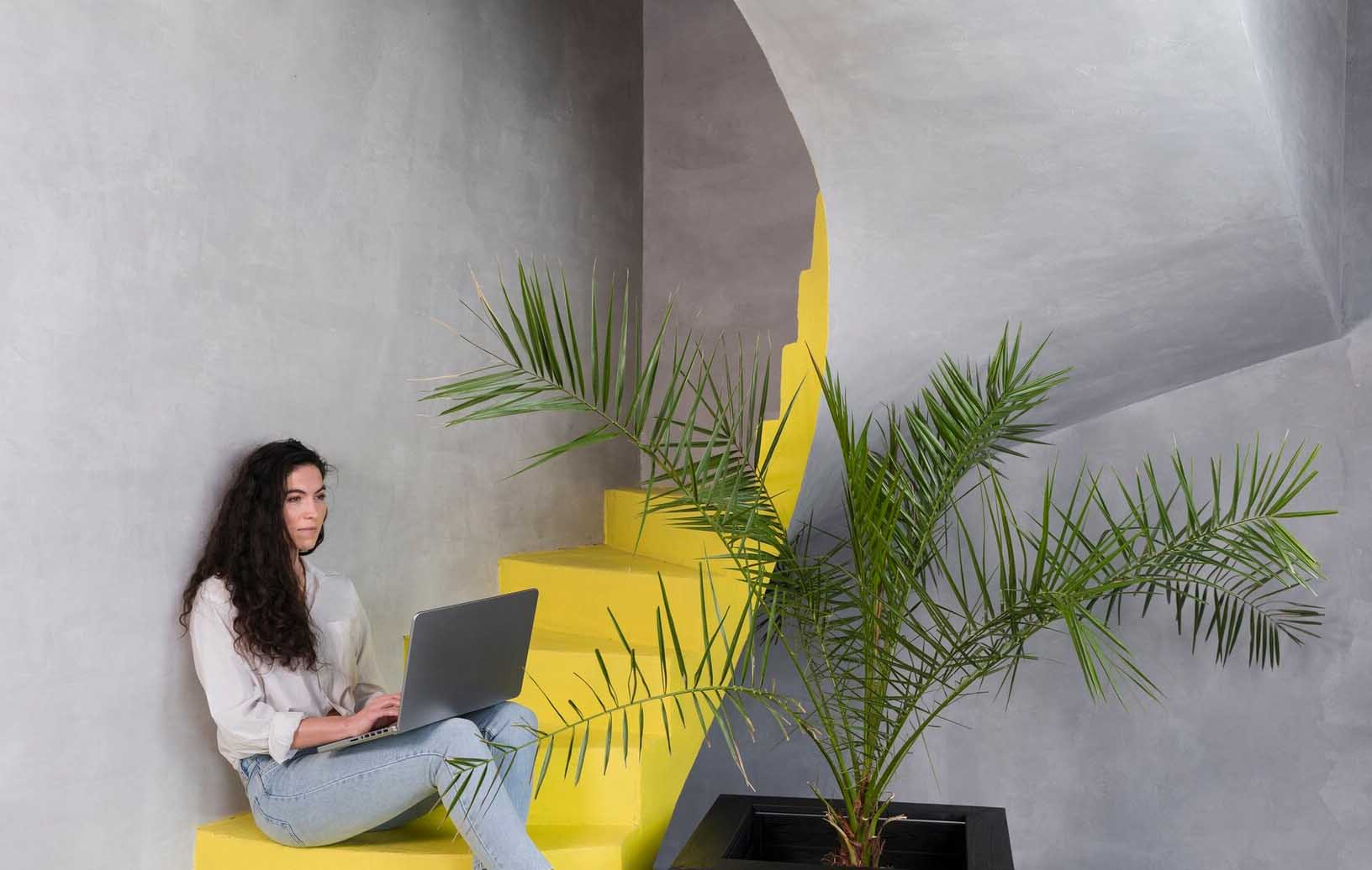 Mulher no laptop em ambiente com escada amarela e planta, com cores na decoração que inspiram home office