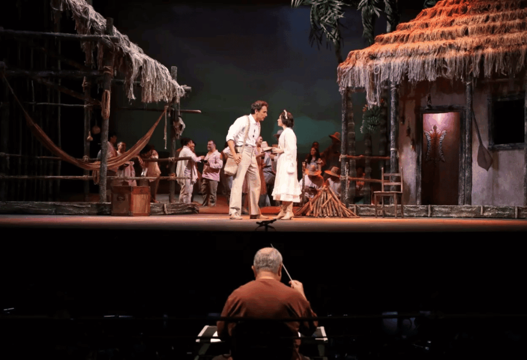 Imagem de apresentação no Festival Amazonas de Ópera, com cenário cenográfico inspirado em ambiente regional e iluminação marcada por tons quentes de âmbar, dourado e marrom.