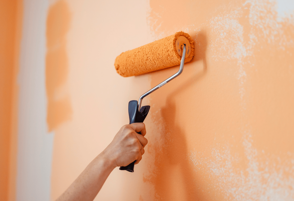 Pessoa aplicando tinta laranja na parede com rolo de pintura.